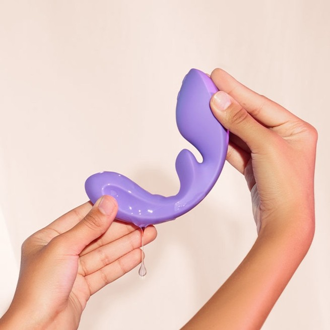 Hands holding a purple Flow, Bedroom Toy Vibrator by Liberator with water on it against a white background.