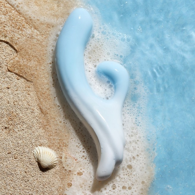 A blue Osurging Rabit Vibrator by Liberator laying in sand next to water.
